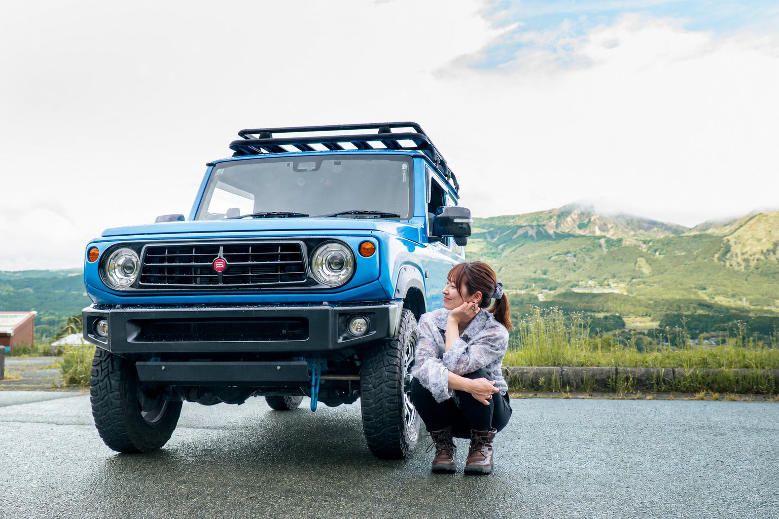阿蘇の山々を背景に、ジムニーのそばでしゃがむやよいさん