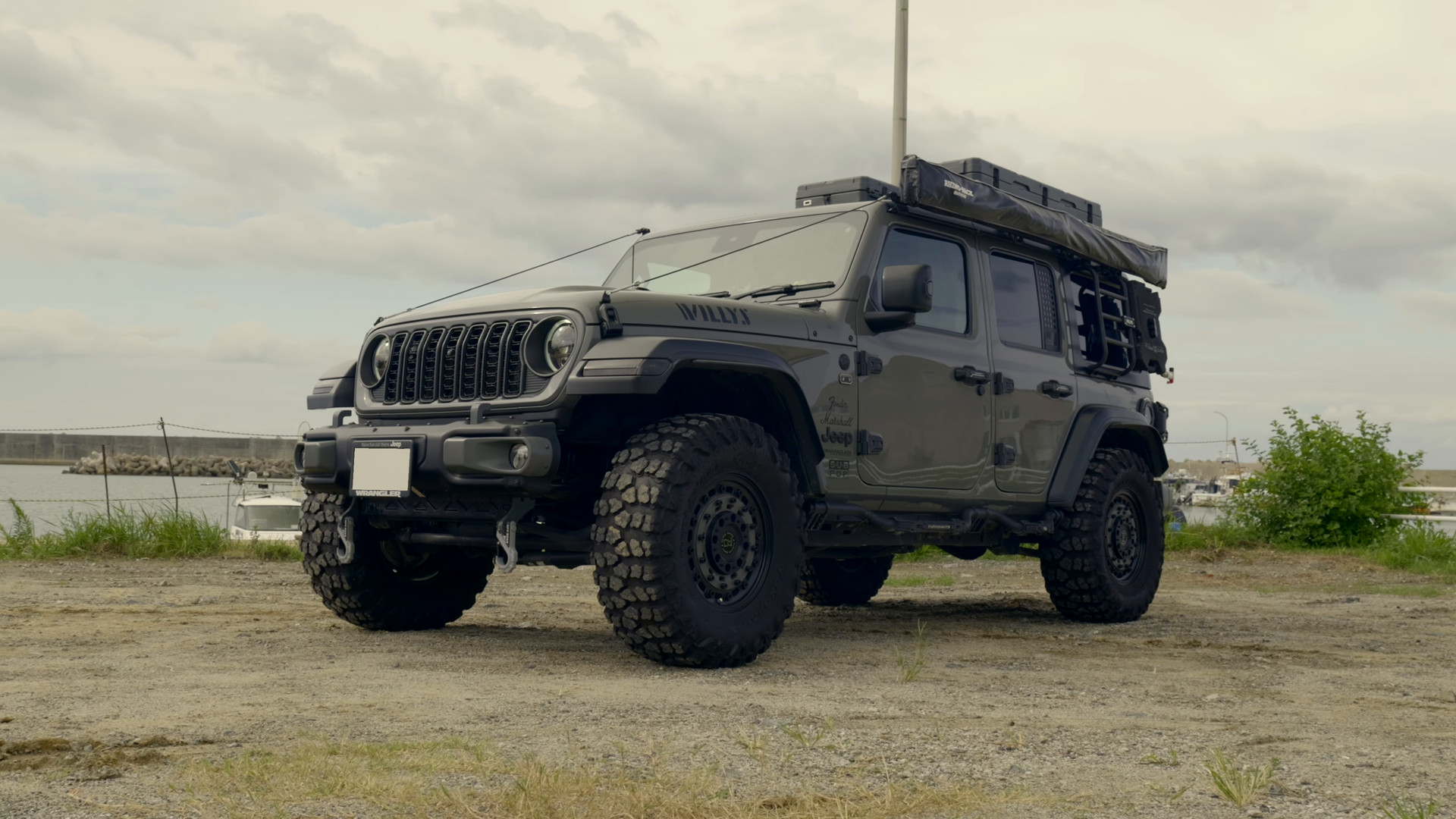 港に停まる、ルーフラックと大型タイヤを備えたグレーのJeepラングラー